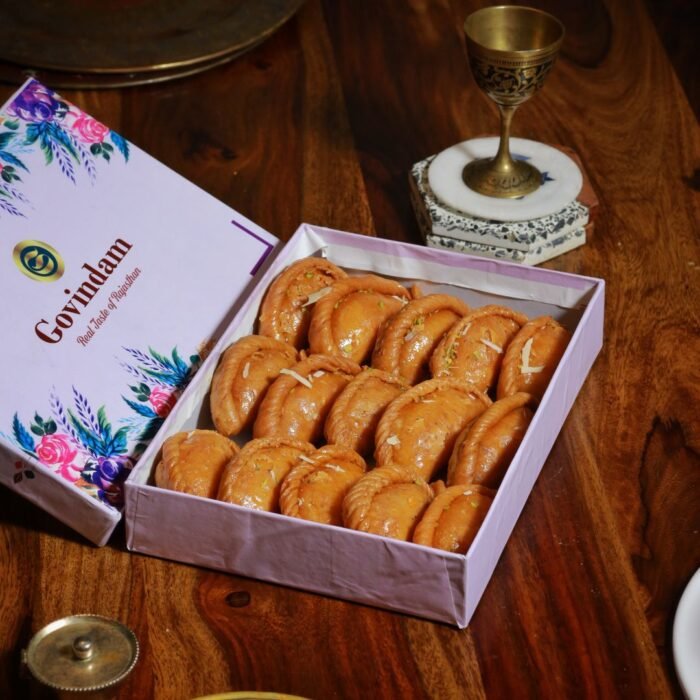 Golden-brown crescent-shaped gujiyas with intricate edges, dusted with powdered sugar on silver platter