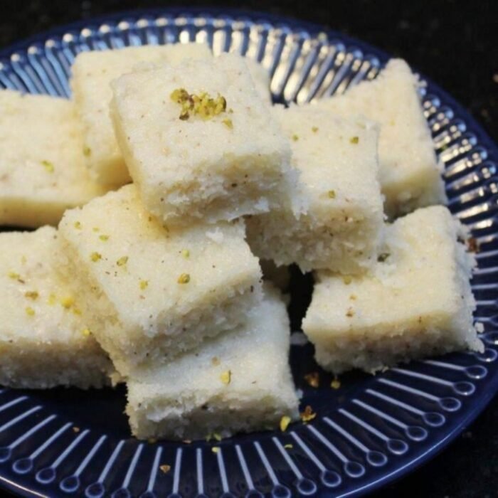 Square pieces of white coconut barfi garnished with slivered almonds on silver platter